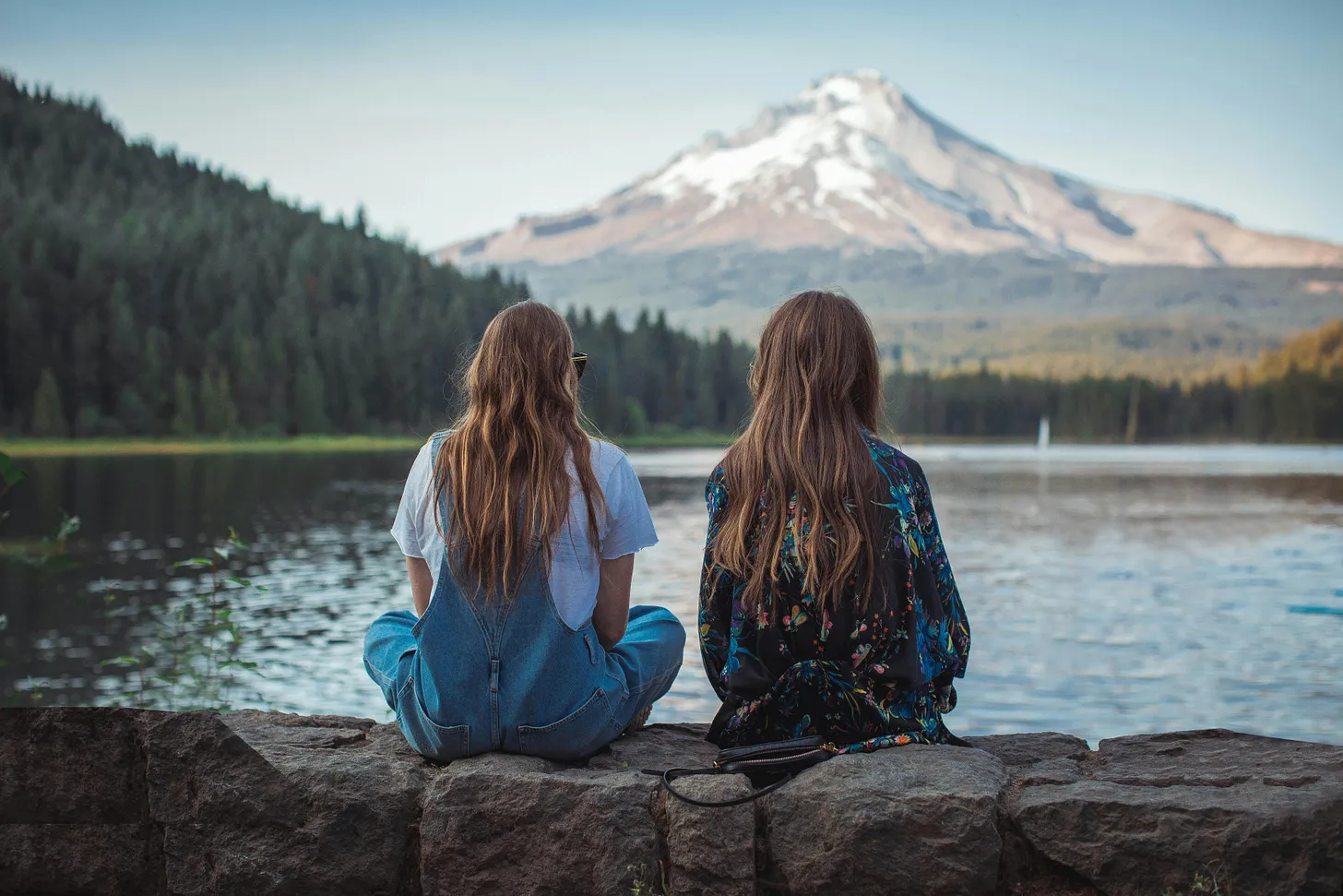 Two women sitting on a rock facing on body of water and mountain