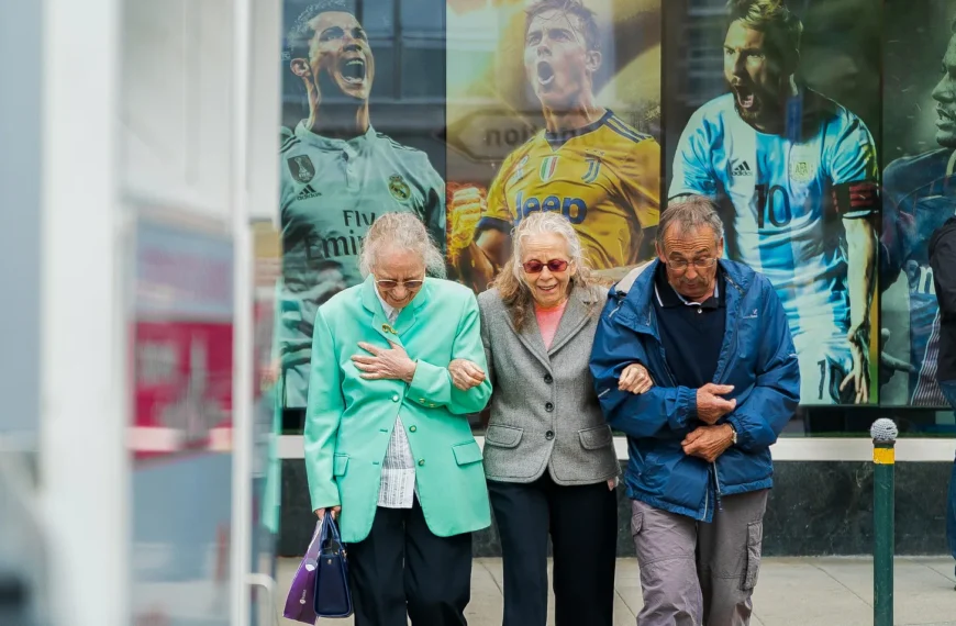 Three elderly people walking, facing the photographer, having a large poster behind them with three soccer players, who are young men, shouting out their victory