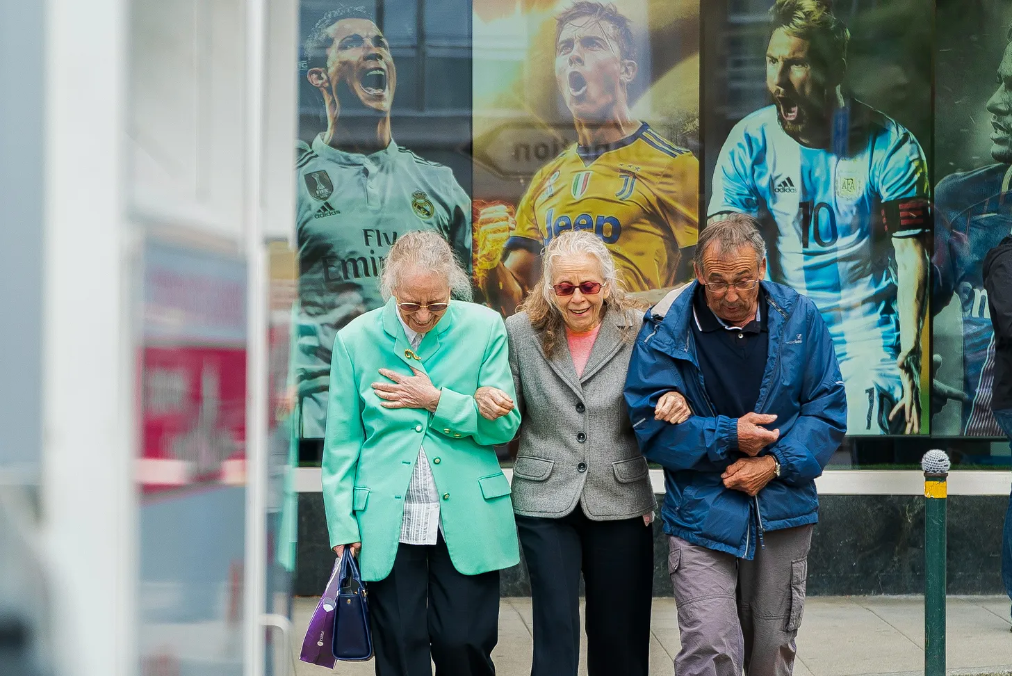 Three elderly people walking, facing the photographer, having a large poster behind them with three soccer players, who are young men, shouting out their victory