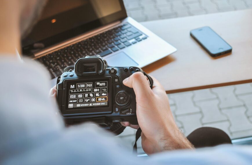 Man holding digital camera, with computer and mobile phone on the table in front of him