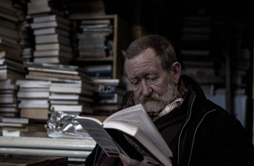 A man is reading a book, with piles of other books behind him