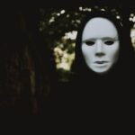 A person covered in black, wearing a ghostly white mask, in dark, outside surroundings with a tree right behind