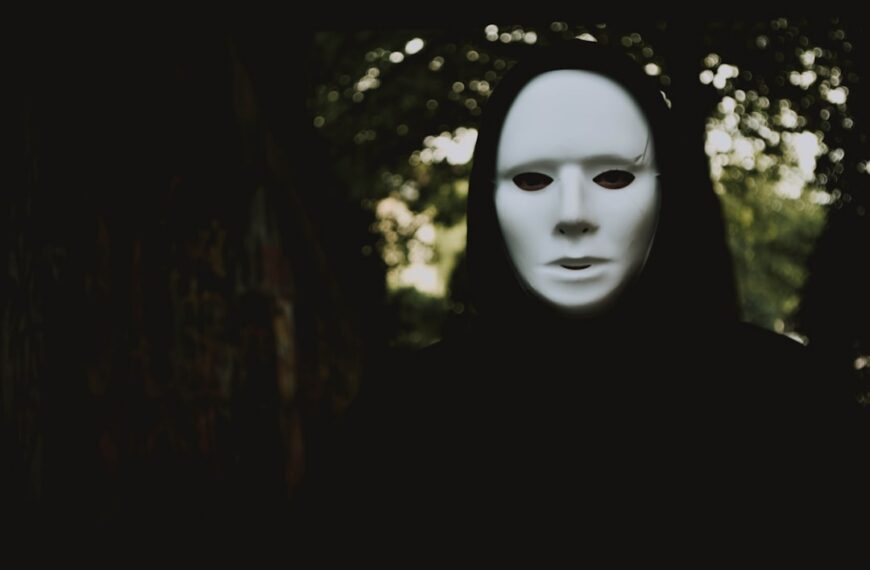 A person covered in black, wearing a ghostly white mask, in dark, outside surroundings with a tree right behind