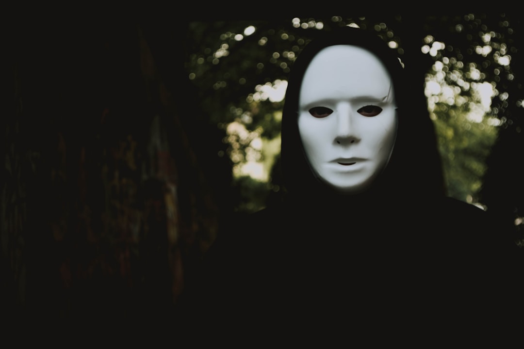 A person covered in black, wearing a ghostly white mask, in dark, outside surroundings with a tree right behind