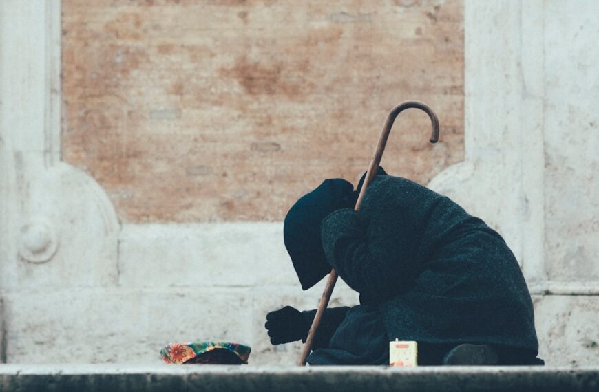 A beggar, sitting on the ground, covered in black, with a hood, in front of a wall