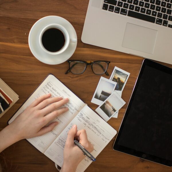 A person is making handwritten notes in a notebook, having books, coffee, a computer, and glasses on the table around the notbook