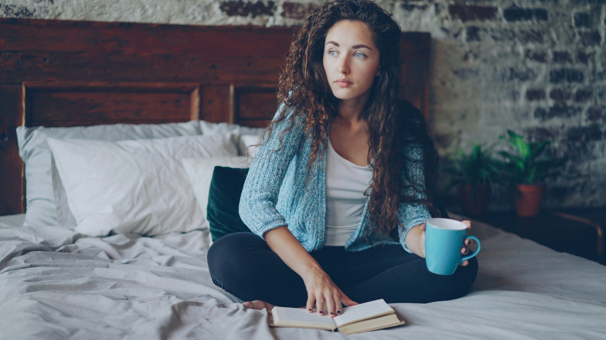 A woman sitting on a bed with an open book in front of her, and a cup in her hand. She looks thoughtful