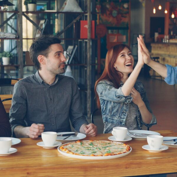 Three people at a table, with a pizza in front of them, and one standing, making a high five with one of those who are siting