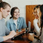 Four people, young adults, sitting around a small but tall table, close to each other, smiling. One is using a tablet computer, another holding a coffee cup, a third has a laptop computer in front of her on the table