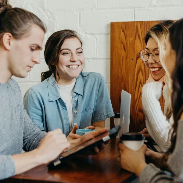 Four people, young adults, sitting around a small but tall table, close to each other, smiling. One is using a tablet computer, another holding a coffee cup, a third has a laptop computer in front of her on the table