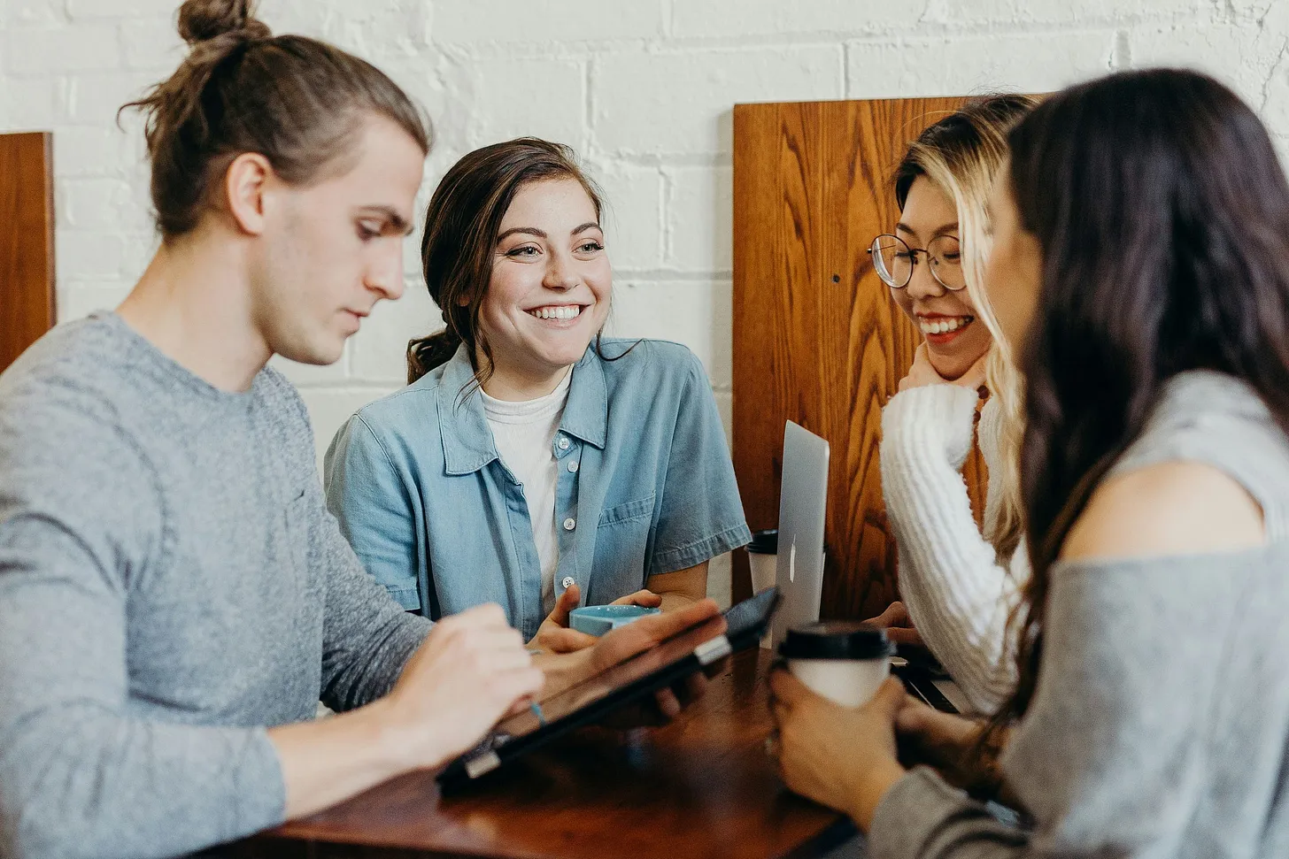Four people, young adults, sitting around a small but tall table, close to each other, smiling. One is using a tablet computer, another holding a coffee cup, a third has a laptop computer in front of her on the table
